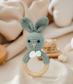 “Flatlay on soft neutral cotton cloth in a minimalist baby nursery. Includes natural props like wooden toys, muslin swaddles, and pacifiers. Calm daylight, warm pastel colors like beige, cream, and soft mint. Eco-style setting with natural wood and handmade baby accessories. Perfect for showcasing a small crochet teether with wooden beads.”