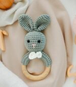 “Flatlay on soft neutral cotton cloth in a minimalist baby nursery. Includes natural props like wooden toys, muslin swaddles, and pacifiers. Calm daylight, warm pastel colors like beige, cream, and soft mint. Eco-style setting with natural wood and handmade baby accessories. Perfect for showcasing a small crochet teether with wooden beads.”