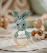 “Flatlay on soft neutral cotton cloth in a minimalist baby nursery. Includes natural props like wooden toys, muslin swaddles, and pacifiers. Calm daylight, warm pastel colors like beige, cream, and soft mint. Eco-style setting with natural wood and handmade baby accessories. Perfect for showcasing a small crochet teether with wooden beads.”